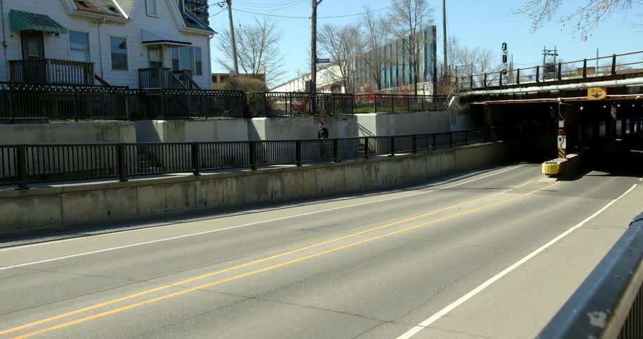 Teenager skateboards on the sidewalk along a road under bridge.