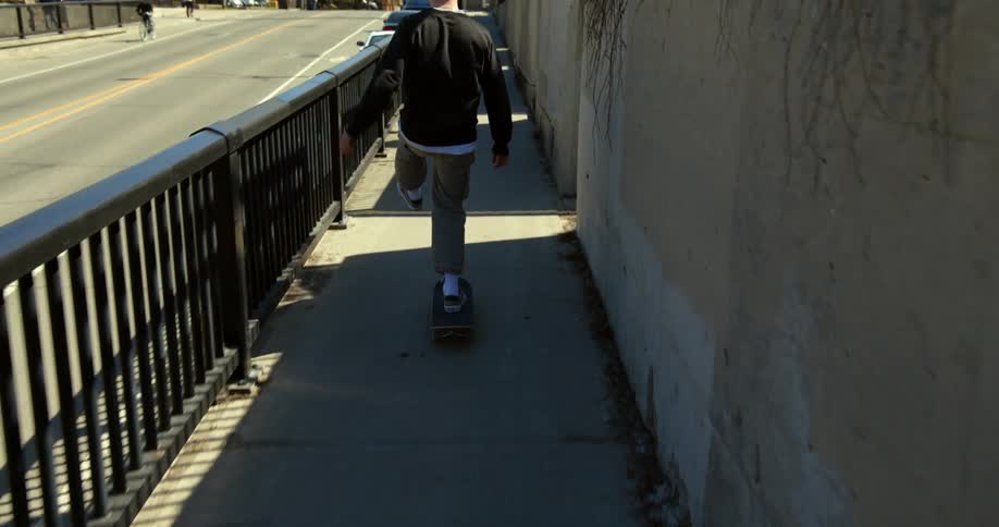 Teenager skateboards on the sidewalk along a road under bridge.