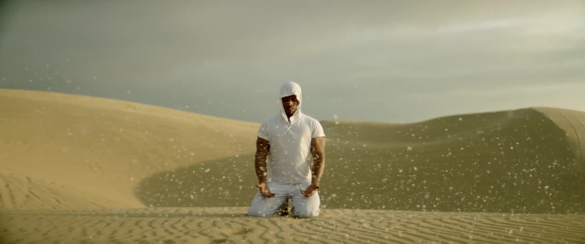 Man kneels praying in an empty desert as sand blows past him
