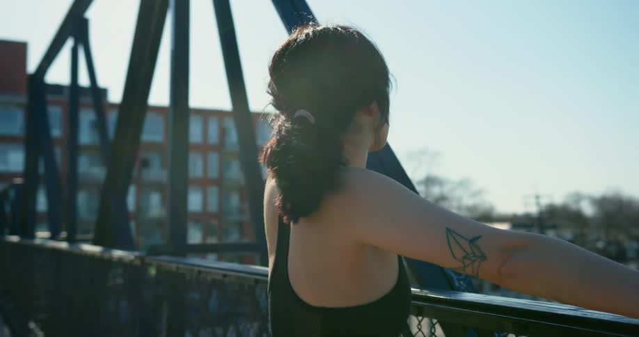 A female jogger stretches and catches her breath on a bridge over train tracks.