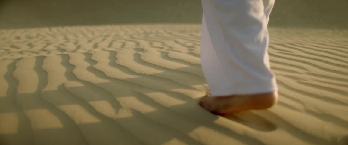 A barefoot man wearing white pants walks on sand