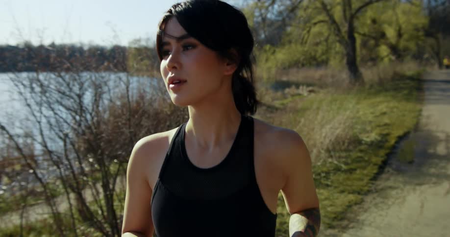 Asian woman jogs along a trail by a lake with city in background.