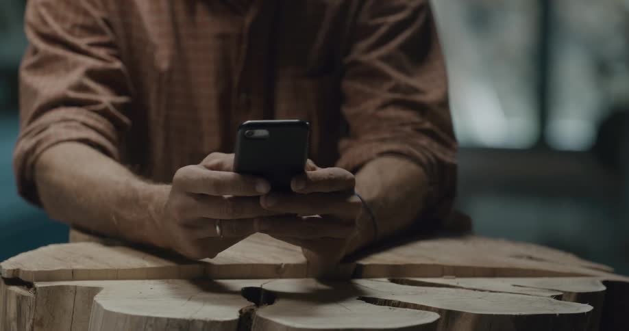 A man leaning on a tree trunk he is carving texts on his cell phone