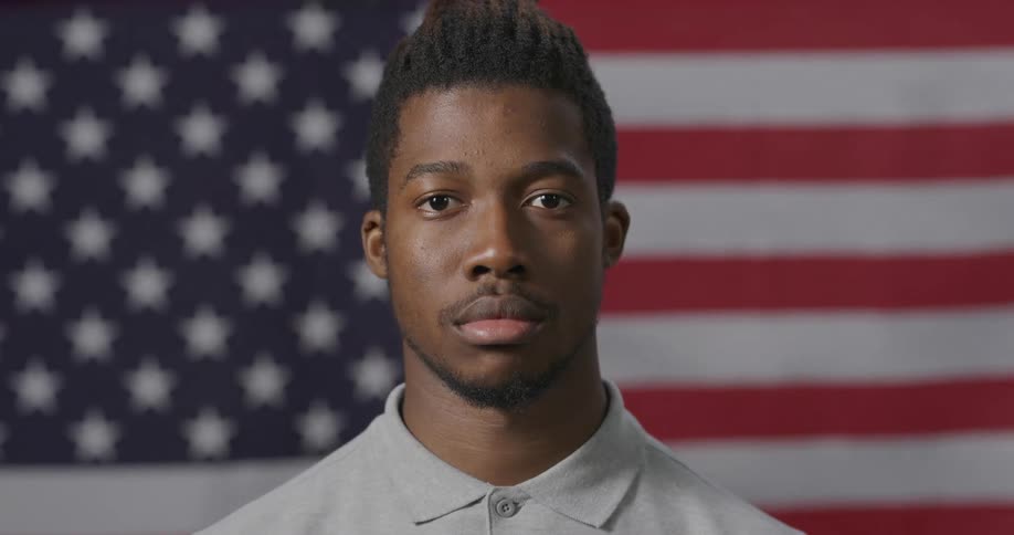 Black man with a grey polo shirt standing in front of the american flag faces the camera with eye contact