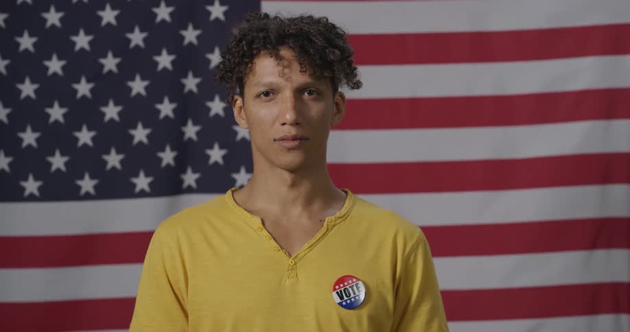 A man stands in front of a hanging american flag with a pin that says to vote