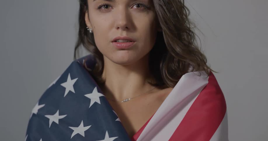 A young woman stares straight ahead as she slow wraps herself in an american flag