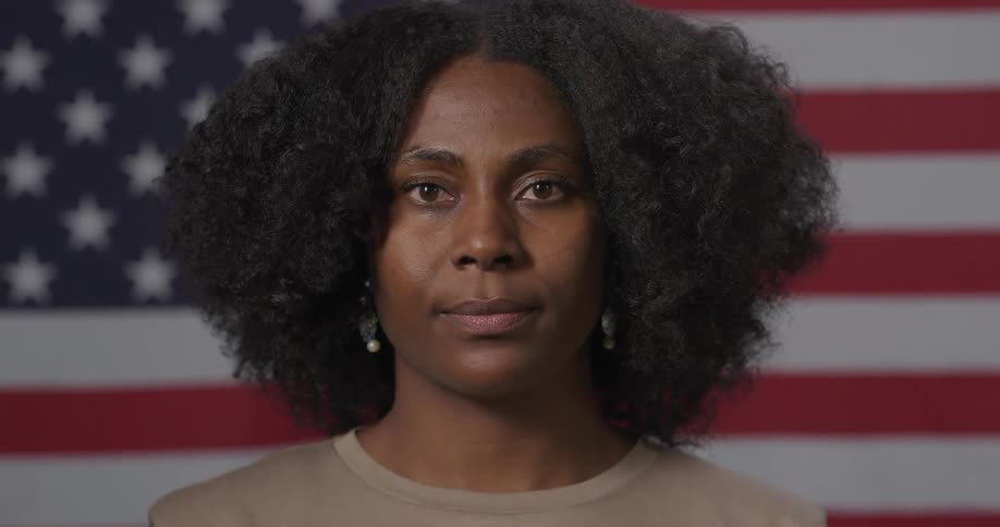 A black woman looks into the camera while standing in front of a fully extended american flag