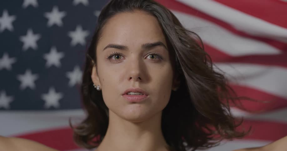 A young woman looks forward has she holds up an american flag stretched behind her head