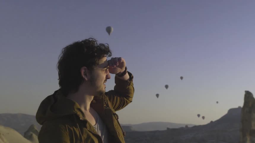 A man in a brown jacket looks out at hot air balloons