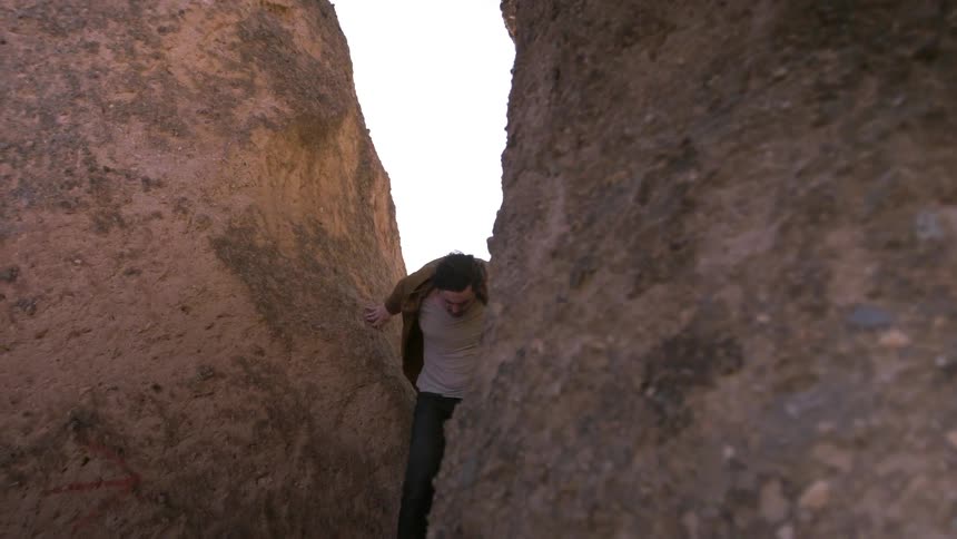 A man in a brown jacket climbs between two rocks