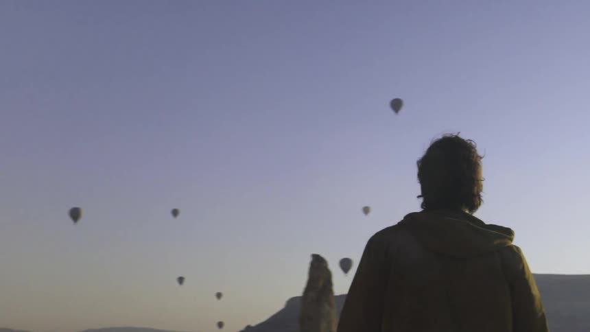 A man stands outside staring at hot air balloons all over the sky