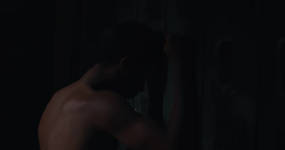In a darkened locker room a muscular young man hits his fist against a closed locker