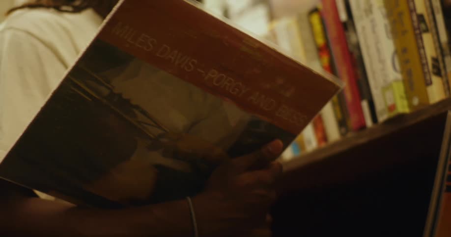Black man with braided hair glasses and a white t shirt looks through a magazine next to a bookshelf