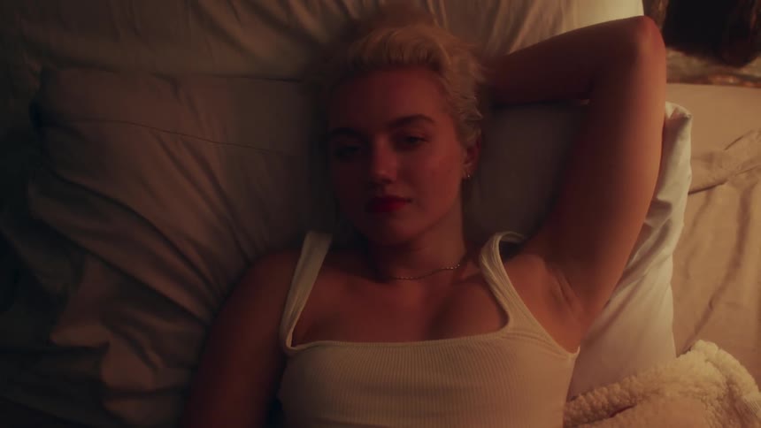 Blonde woman with white undershirt lying in bed looks at the camera in bedroom