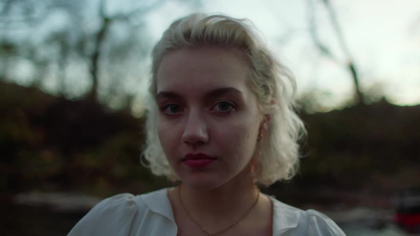 Blonde woman with a white shirt faces the camera with eye contact by a stream