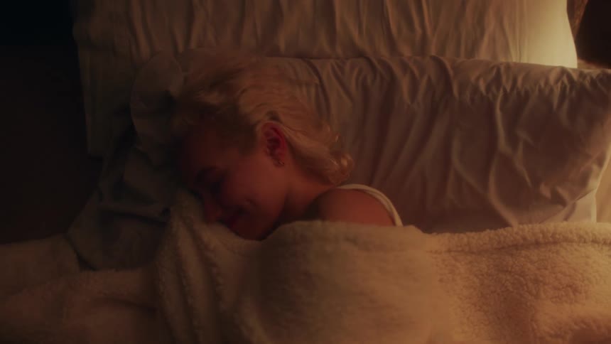 Blonde woman lying down in bed looks at the camera and smiles in bedroom at night