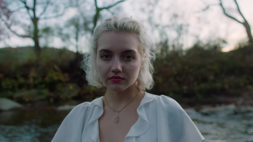 Blonde woman with makeup a necklace and a white shirt faces the camera next to a stream at dusk