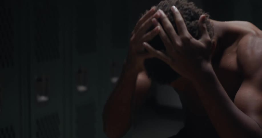 A teenage boy in a locker room rubs his hands together and puts his face and head in his hands