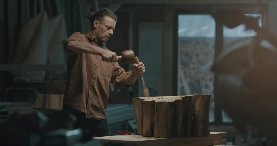 A man with a chisel sculpting a piece of wood on a table
