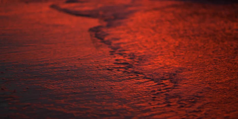 Waves sweep over the sand on a beach soaked in a reddish light