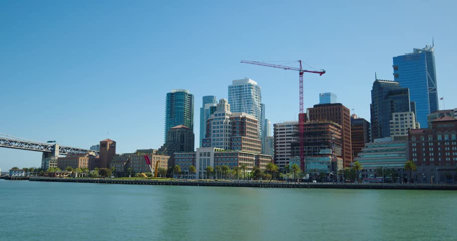 A shoreline with trees buildings construction cranes and pedestrians with the sky in the background