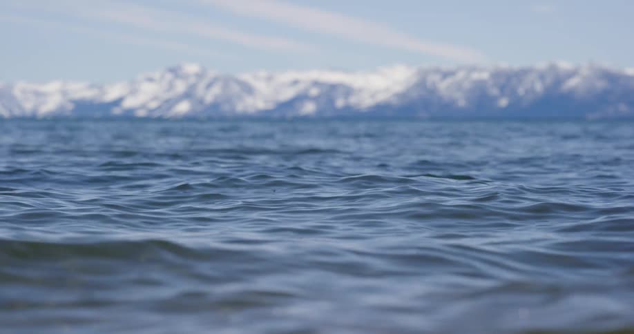 Small waves flow across surface of water paralleling the snow covered mountains on the horizon under a cloudy sky