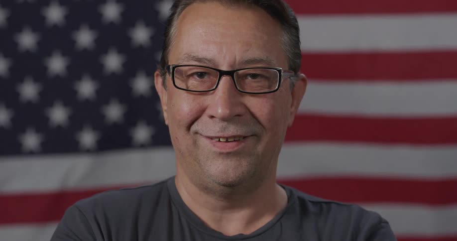 Middle aged man with glasses raises his head looks at the camera with eye contact and smiles in front of the american flag