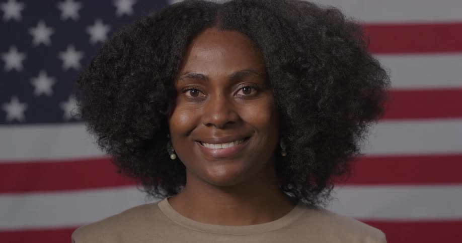 Black woman with curly hair faces the camera with eye contact and smiles warmly in front of the american flag