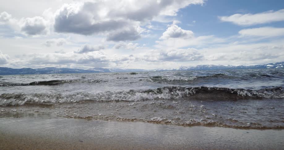 Waves crash onto beach under cloudy sky while snow covers mountains on horizon