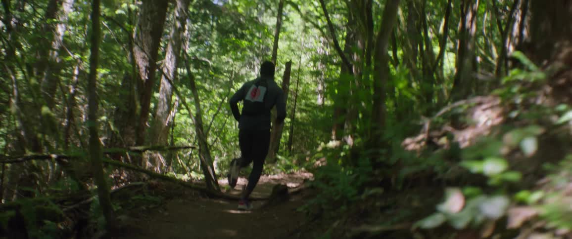 A male athlete is jogging up a dirt trail in a forest during the day