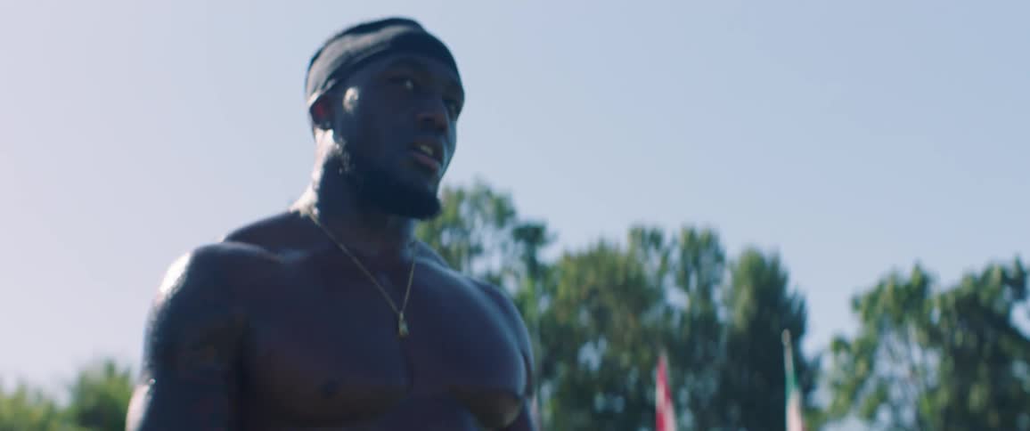 A shirtless man breathes heavily and sweats while standing on an athletic field