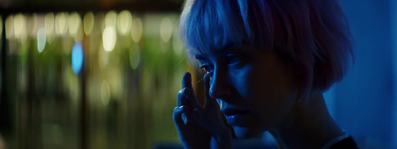 A crying young woman wipes the tears from beneath her eye with a finger as she stands alone in a dimly lit area