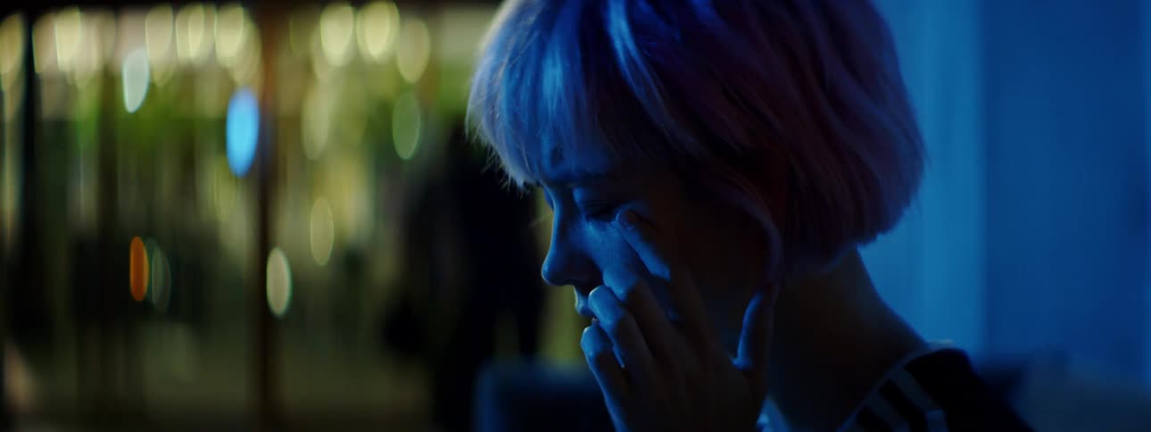 A teenage girl sitting on a couch indoors cries and wipes her eyes as other people walk past