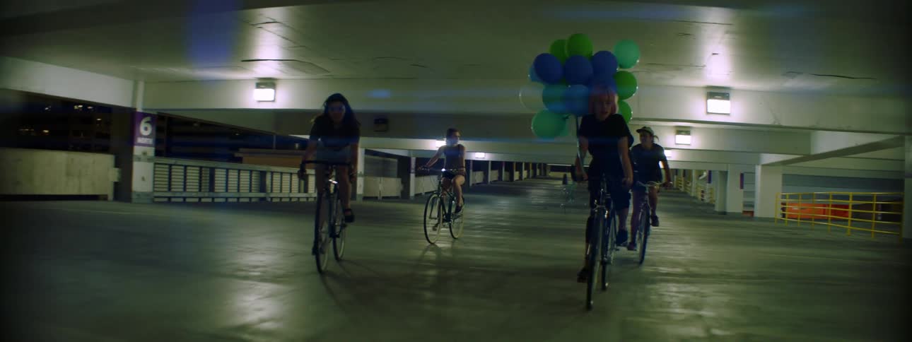 Four teenage girls one with a big bouquet of balloons ride their bikes through a parking garage