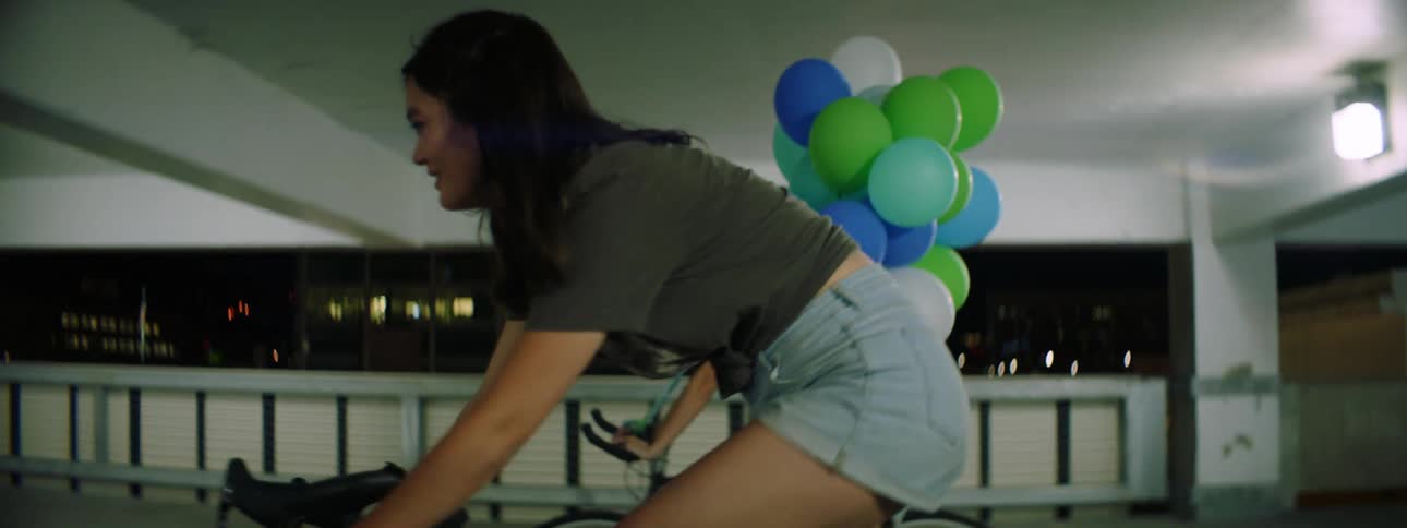 Two teenagers one carrying balloons bike through a parking garage at night