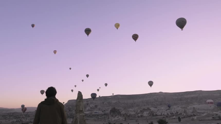 A man is standing outside with many hot air balloons in the sky