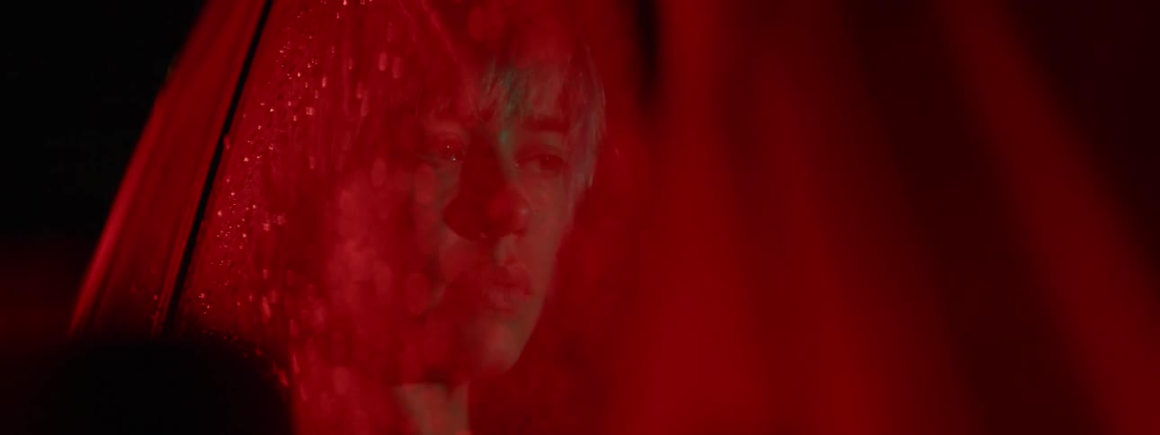 Young woman sits on the passenger seat of a car looking out of the window covered in water droplets under a red light