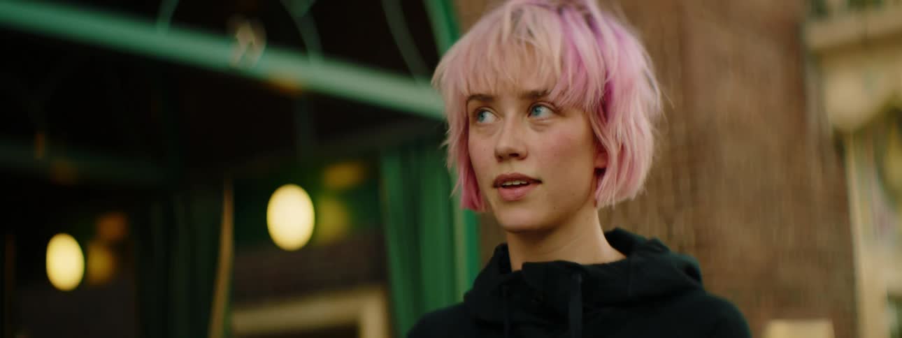 A woman with pink hair talking to a man then walking away with a building in the background