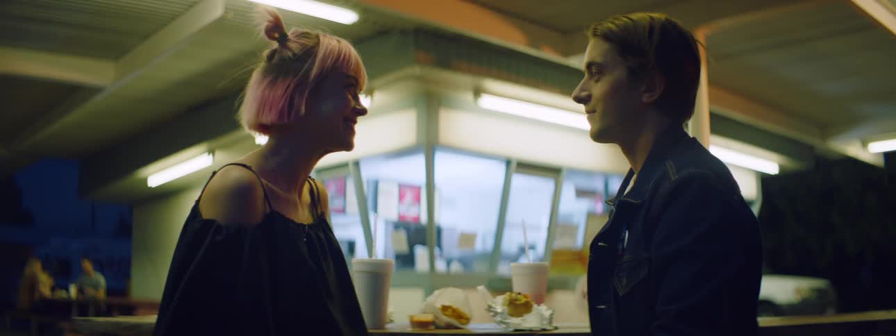 A teenage girl takes a drink at a fast food restaurant and a teenage boy leans forwards and kisses her
