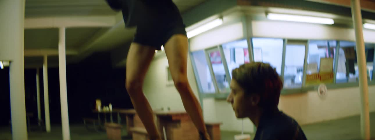 A woman with pink hair dances on top of a table outside a walk up style restaurant as her boyfriend looks away