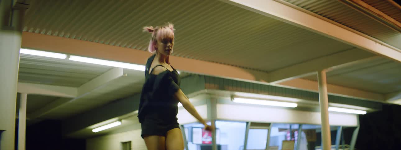 A man sitting outside a restaurant at night tries to eat his food as a young woman with pink hair dances atop his table