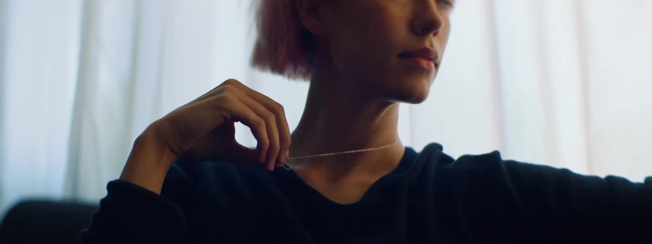 Woman with pink hair sitting on a dark blue couch fiddles with her necklace