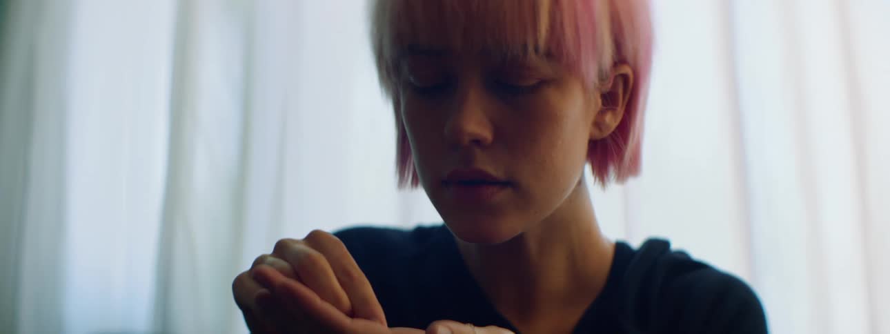 Woman with pink hair sits on a sofa and fiddles with a necklace in a room