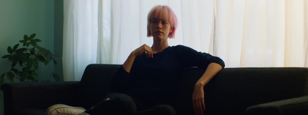 Woman with pink hair sits on a black sofa and looks around in the living room
