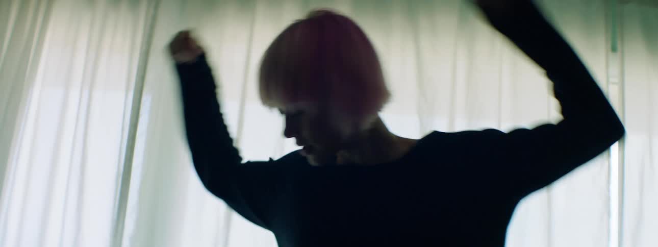 Woman with pink hair dances as she stands on a sofa in front of a window