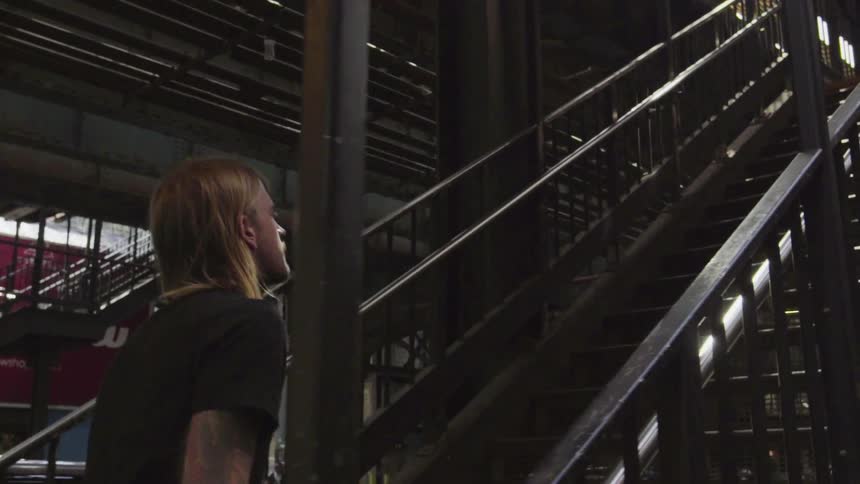 A tattooed man walks up outdoor stairs to a train platform in a city