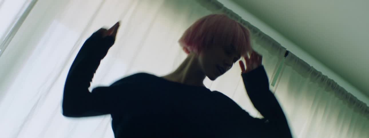 Woman with pink hair and a long sleeve black t shirt dances wildly on a black sofa in a room