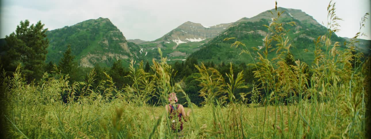 Woman with pink hair and a red backpack walks through tall grass in an area of land covered with mountains
