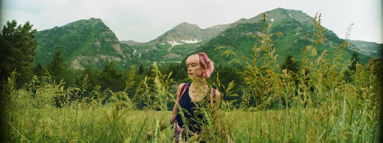 A young woman hikes through a field of tall grass below a mountain ridge on a sunny day