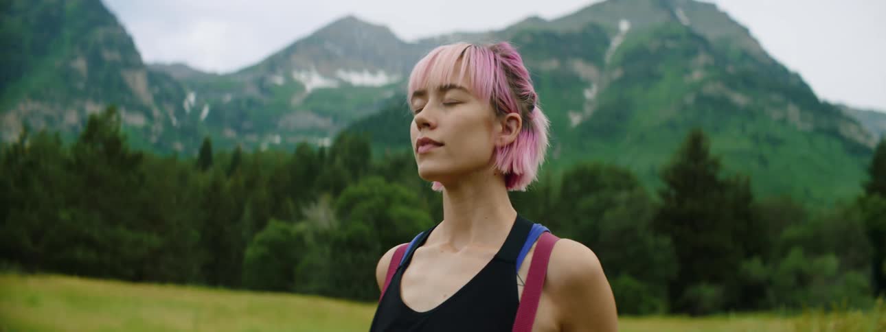 Young woman with pink hair wearing a backpack stands in a grass field next to the mountains